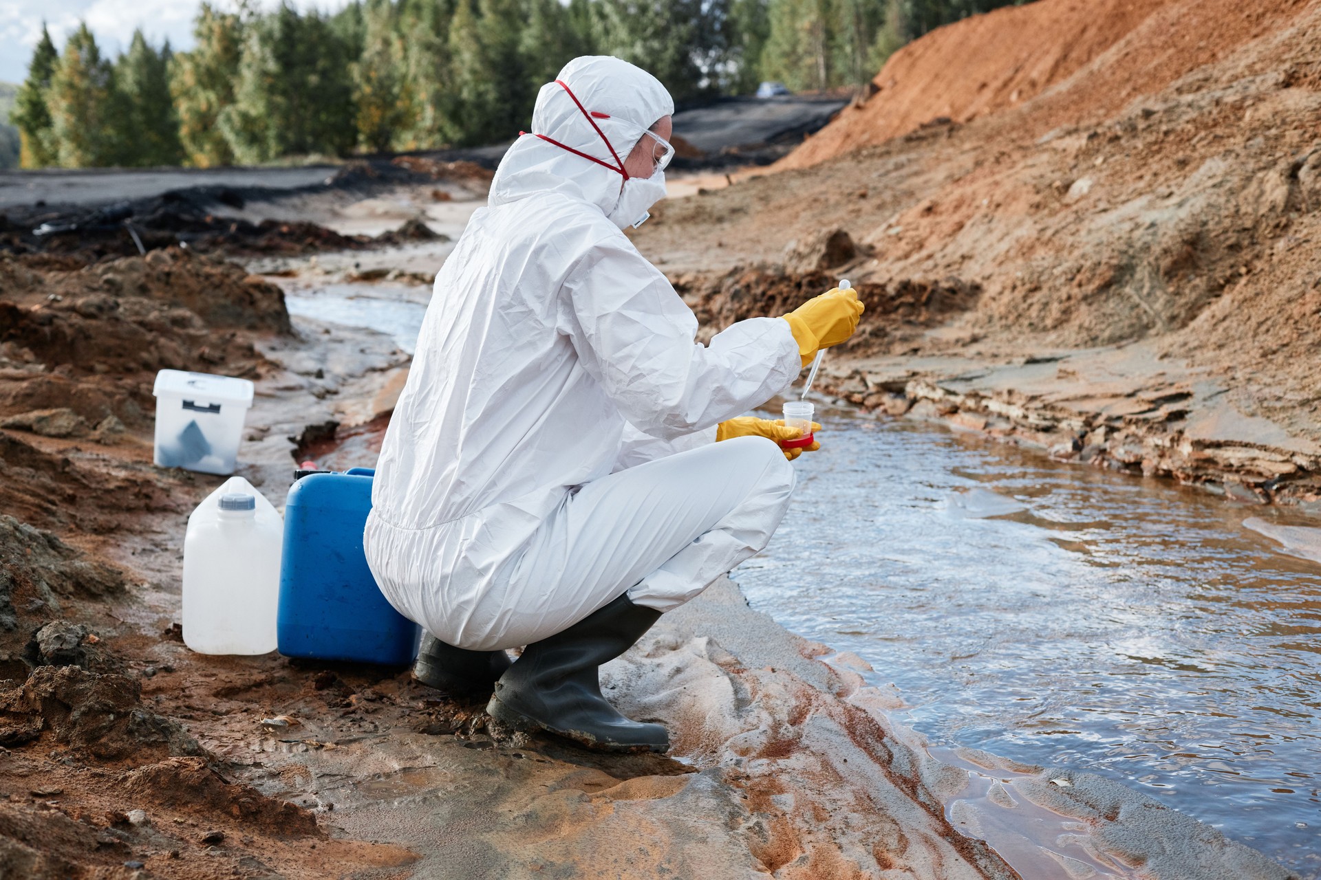 Scientifique terrestre en combinaison biohazmat, masque et gants accroupis à la rivière et déposant un échantillon d’eau dans un récipient